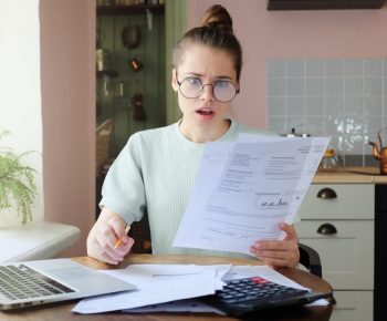 Amazed,Young,Woman,Sitting,At,Kitchen,Table,,Holding,Utility,Bills