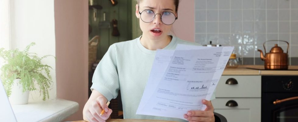 Amazed,Young,Woman,Sitting,At,Kitchen,Table,,Holding,Utility,Bills